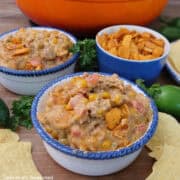 Two white bowls with blue trim filled with a serving of cheesy queso taco dip and a blue bowl filled with coarsely crushed chili cheese Fritos in it.