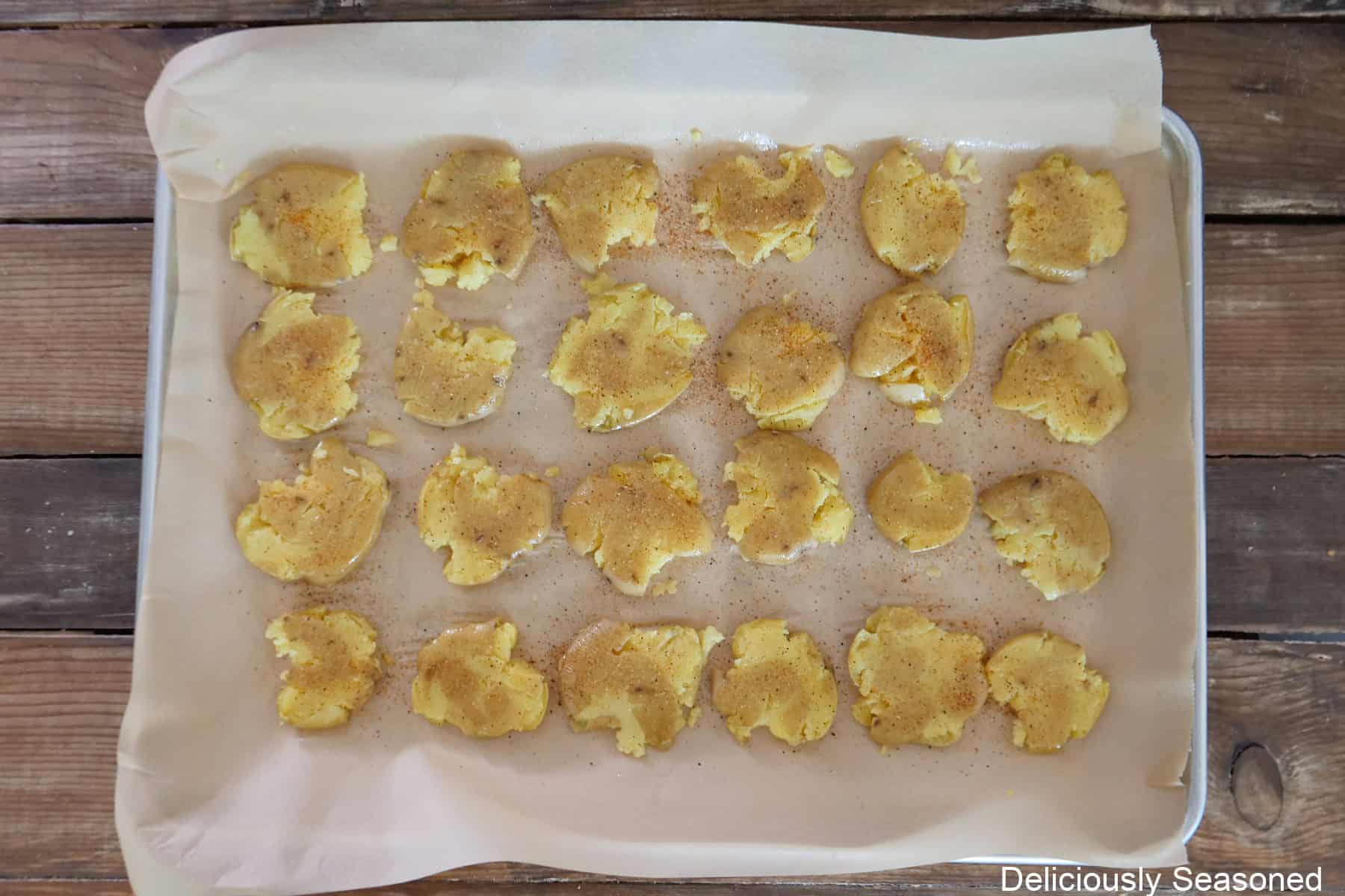 A baking sheet lined with parchment paper with smashed mini potatoes on it seasoned and drizzled with olive oil before being baked.