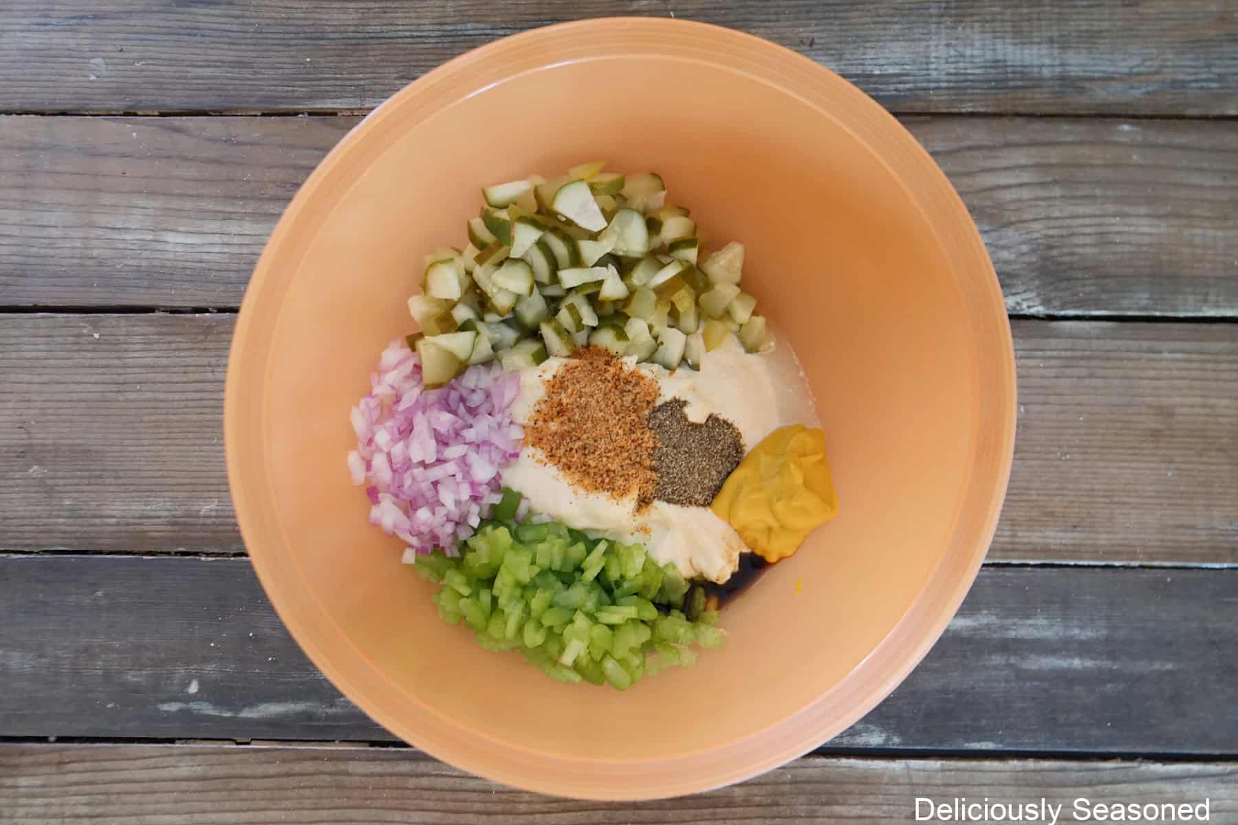 An orange mixing bowl filled with all the creamy dressing, veggies, seasonings, and diced pickles placed in it before being mixed together.