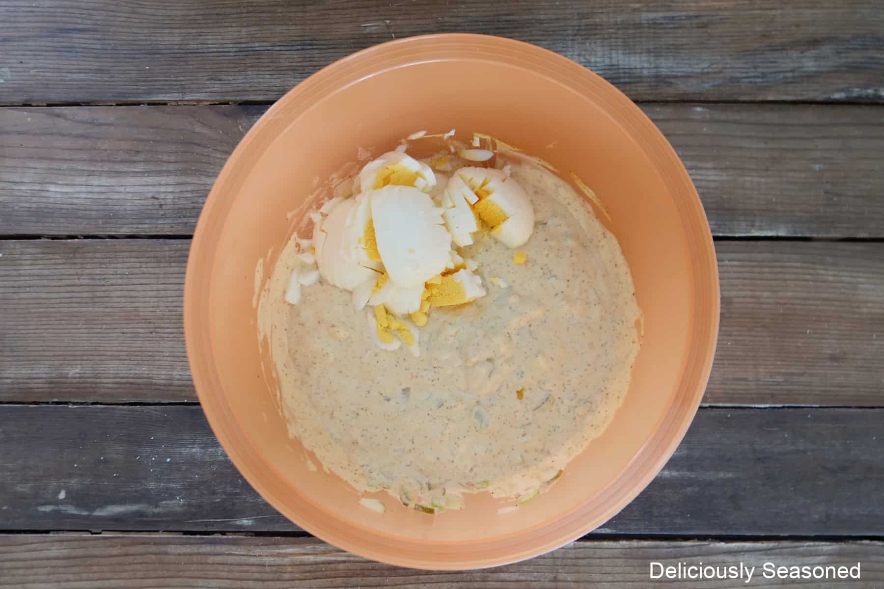 A woos surface with an orange mixing bowl with creamy potato salad dressing with diced boiled eggs added.