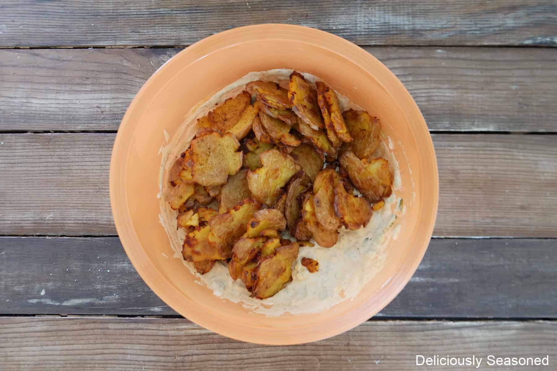 A wood surface with an orange mixing bowl filled with all the potato salad ingredients and baked smashed potatoes added with the creamy dressing.