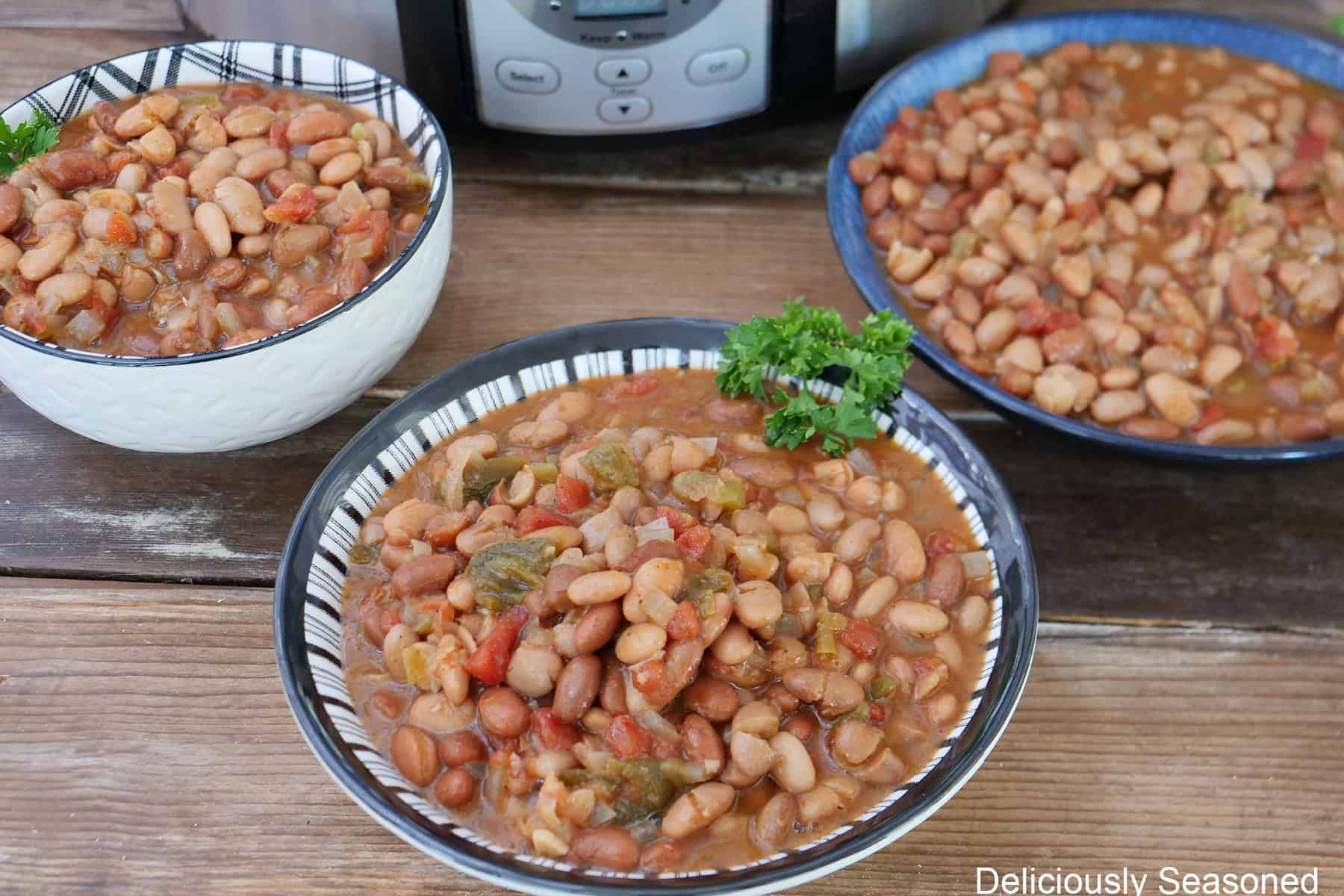 A rustic wood surface with three bowls filled with spicy beans and a crockpot in the background.