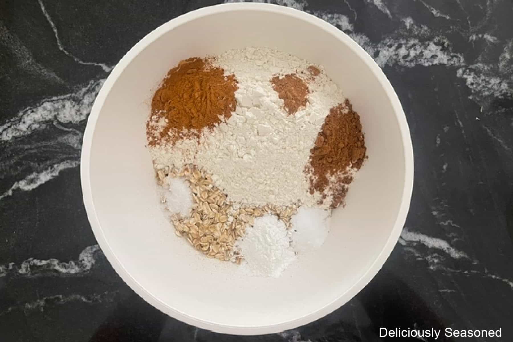 A black countertop with a white mixing bowl filled with dry cookie ingredients before being combined.
