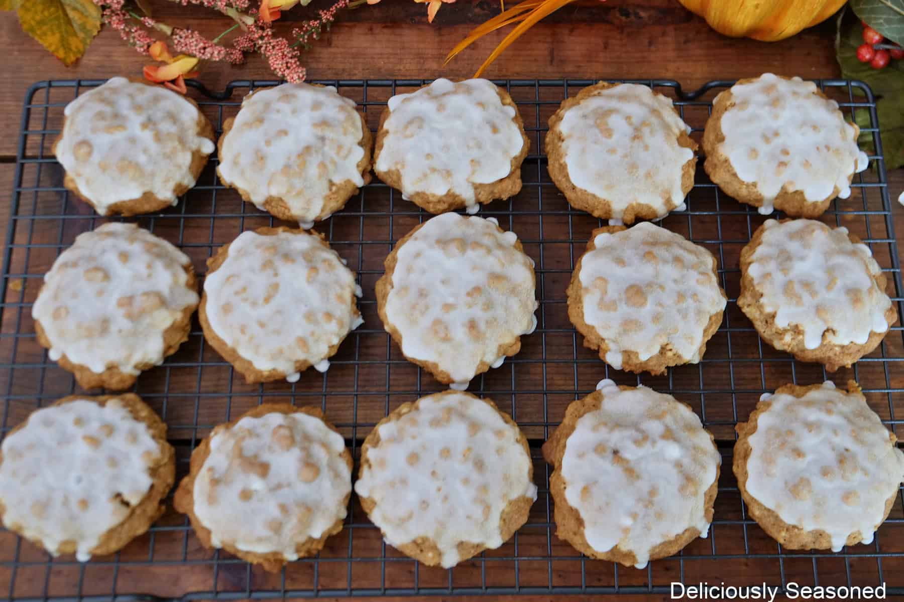 A wire rack with fifteen maple iced pumpkin oatmeal cookies on it with fall foliage in the background.