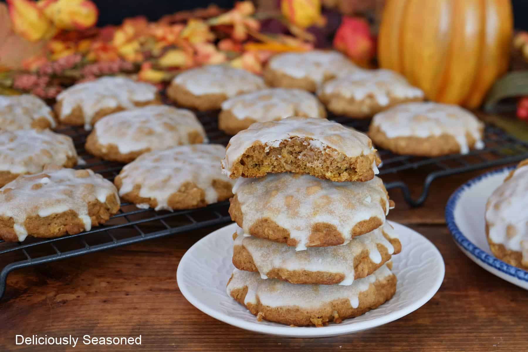 A wood surface with a small white plate with four cookies stacked on it and a wire rack with more cookies in the background along with fall foliage.