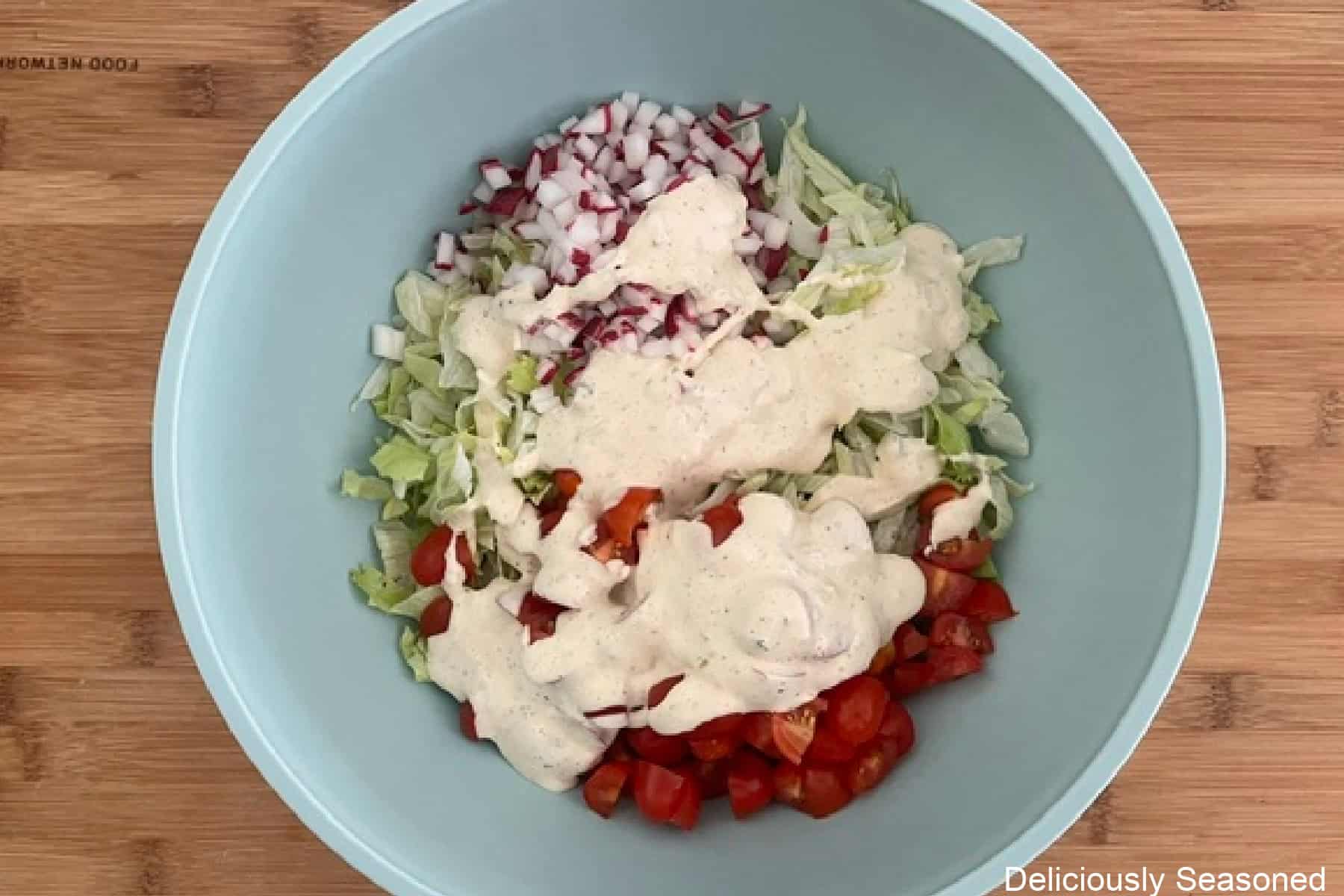 A light blue mixing bowl with shredded lettuce, diced tomatoes and radishes, topped with ranch dressing