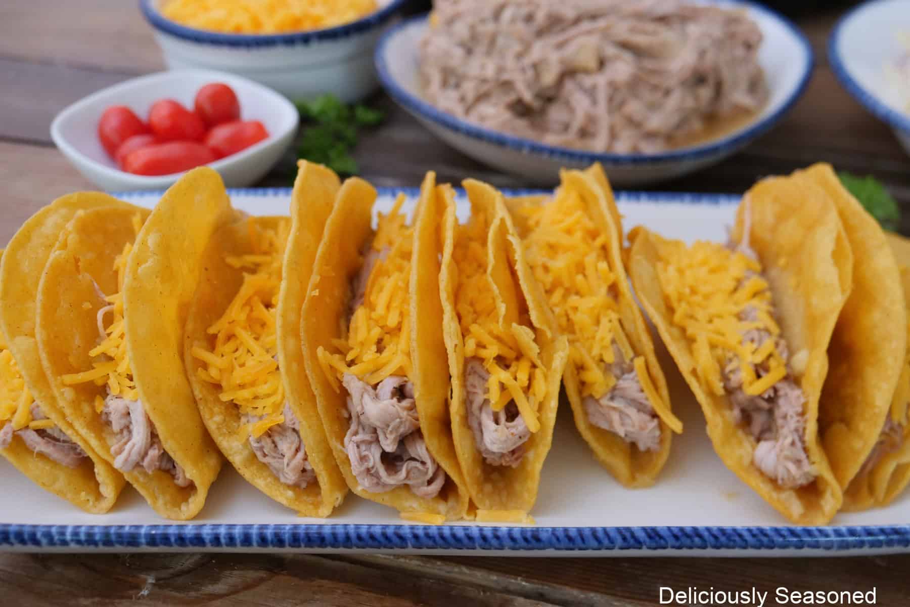 A white oblong plate with blue trim with eight fried corn tortillas filled with shredded pork and shredded cheese.