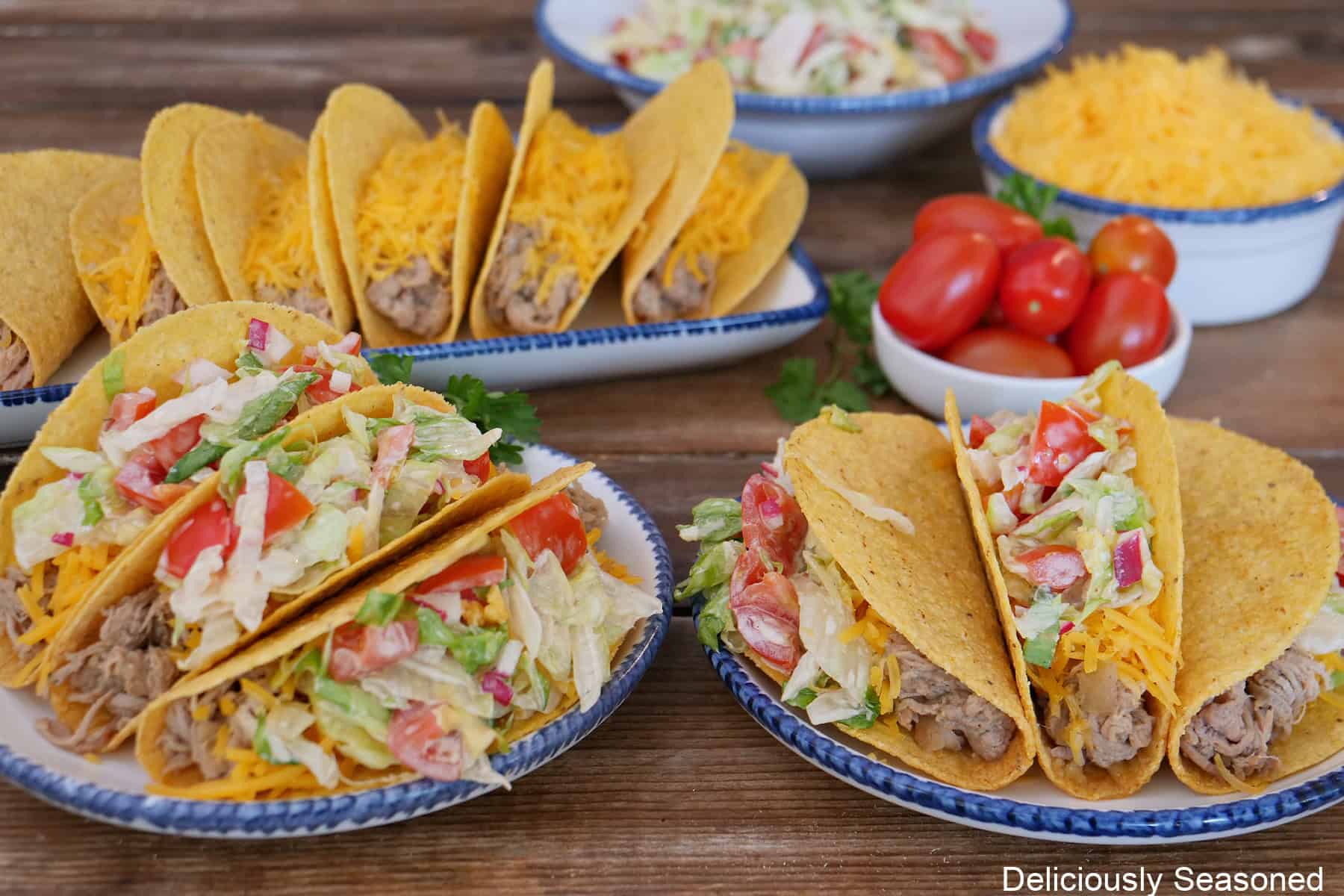 Two round white plates with blue trim with three tacos on each plate, with more tacos in the background.
