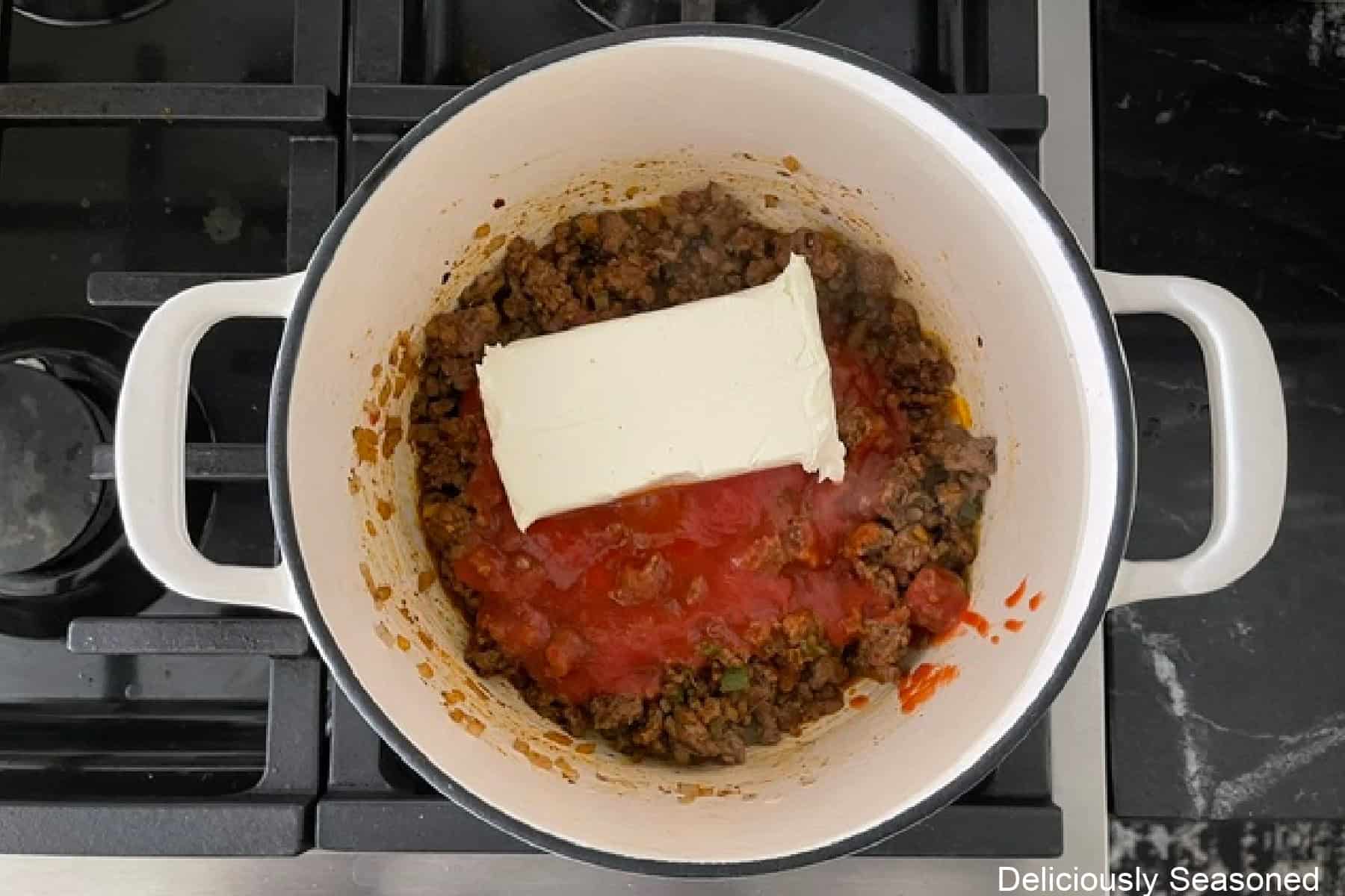 A block of cream cheese and diced tomatoes added to a ground beef mixture in a white cast-iron skillet.